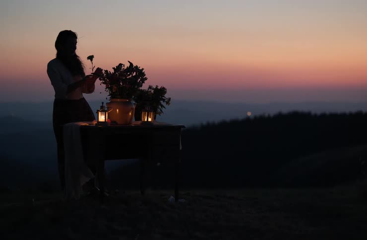 Silhouette of a woman beside a table with candles and flowers against a colorful dusk horizon