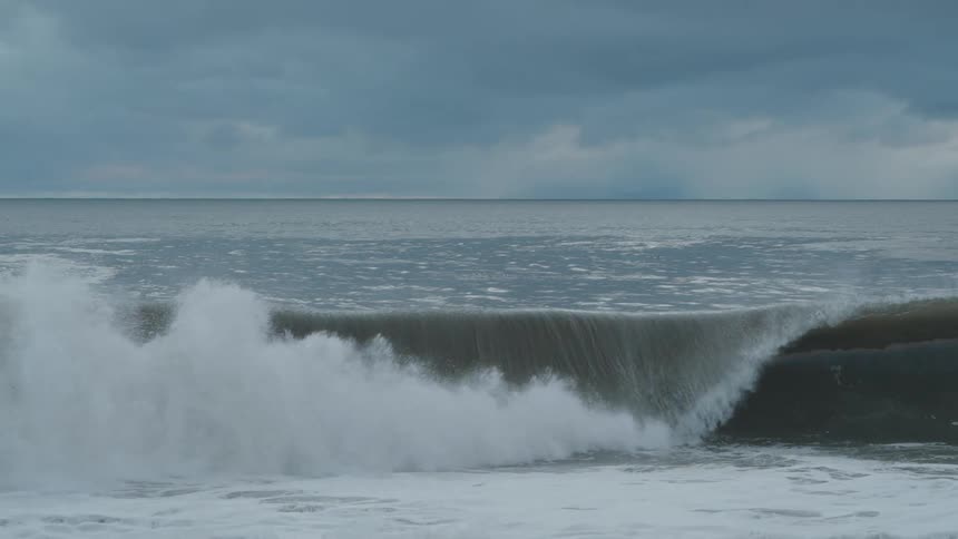 A large dark wave rises and crashes onto the shore creating a thick white spray of foam the scene is dominated by a gray overcast sky and a dark choppy ocean surface suggesting stormy weather