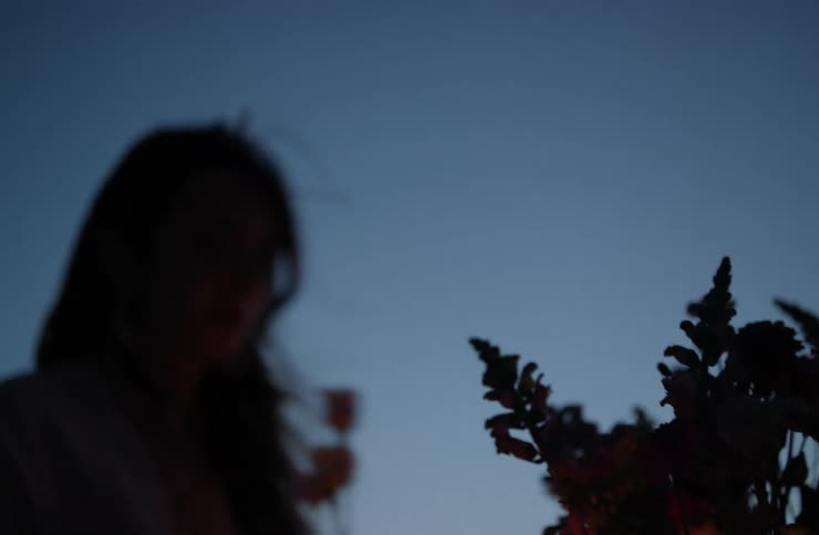 A young woman outdoors at dusk arranges flowers in a vase with a lamp beside her