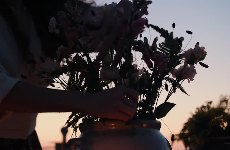 Person arranges flower bouquet in ceramic vase at sunset with softly toned sky