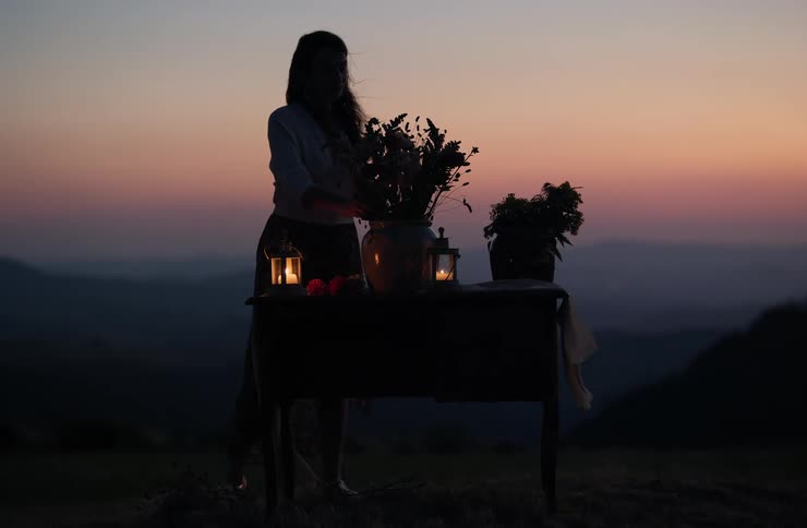 Silhouette of a woman arranging flowers on a table with lit candles against the dusk horizon