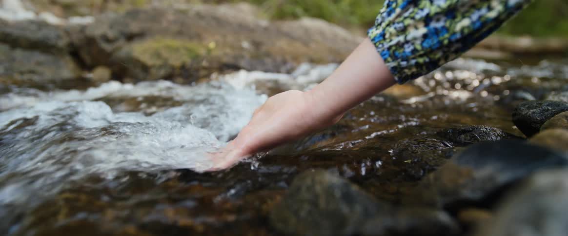 The video is a closeup shot focusing on a womans hand featuring bright pink nail polish gently touching the surface of clear fastflowing water in a stream or small river the water rushes over dark rocks in the foreground creating a natural serene