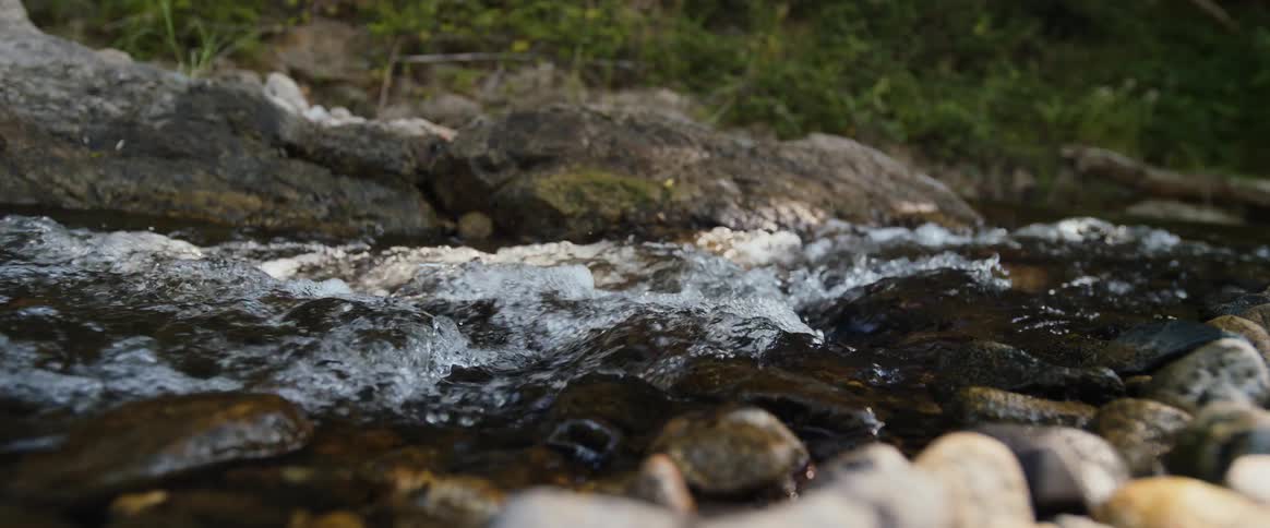 Clear water rushes and bubbles in slow motion over dark smooth rocks in a shallow forest stream creating a peaceful steady flow