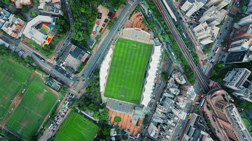 A highangle aerial shot focuses on a modern green soccerfootball stadium tightly integrated into a bustling highdensity city environment the stadium is surrounded by tall residential and commercial buildings major roadways and a clearly visible el