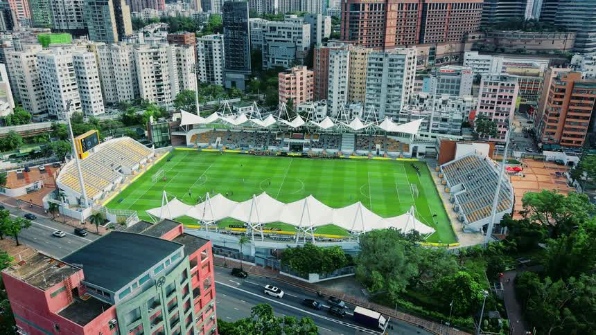 The video is an aerial shot of a football soccer stadium surrounded by dense urban development featuring tall residential and commercial buildings traffic is moving on a multilane road adjacent to the stadium which has a bright green field and disti