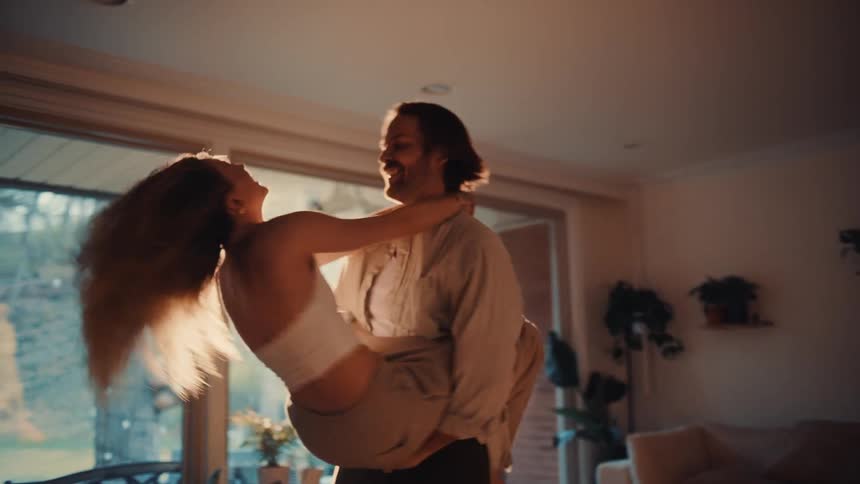 In a sunlit living room a man lifts and spins a woman around in a joyous intimate momentthey are laughing and embracing their movements full of energy and affection