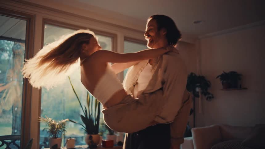 A young man carries a young woman spinning her around in the living room as sunlight streams through the window both of them having fun in excitement