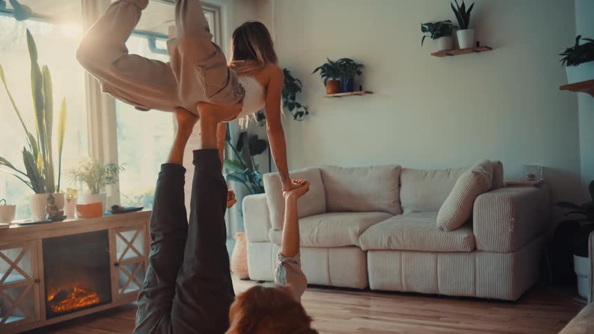 Man lying on the floor lifting a woman with his legs while a dog watches in the sunlit living room