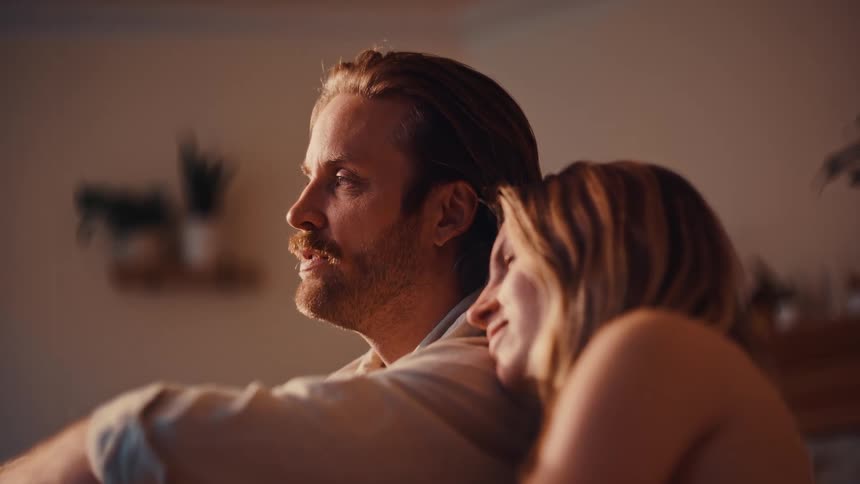 A young woman leans her head on her husbands shoulder indoors as they have a conversation at midday