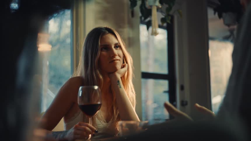 A woman with long blonde hair is seated at a table looking offcamera to her right as if listening to someoneshe is holding a glass of red wine resting her head on her hand and the scene is dimly lit with natural light coming from a large window b