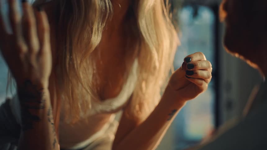 Tattooed hands with black nails clasped together under soft light near a window