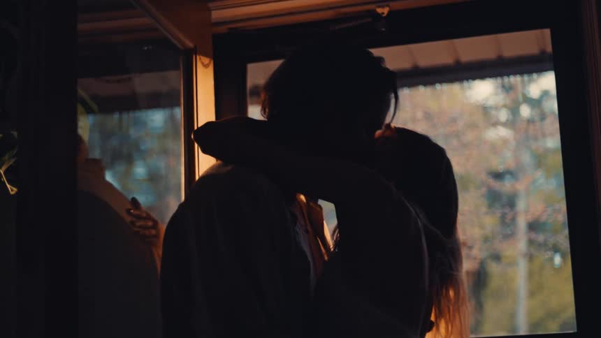 A couple stand indoors hugging and kissing as rain drips from the roof visible through the glass window at midday