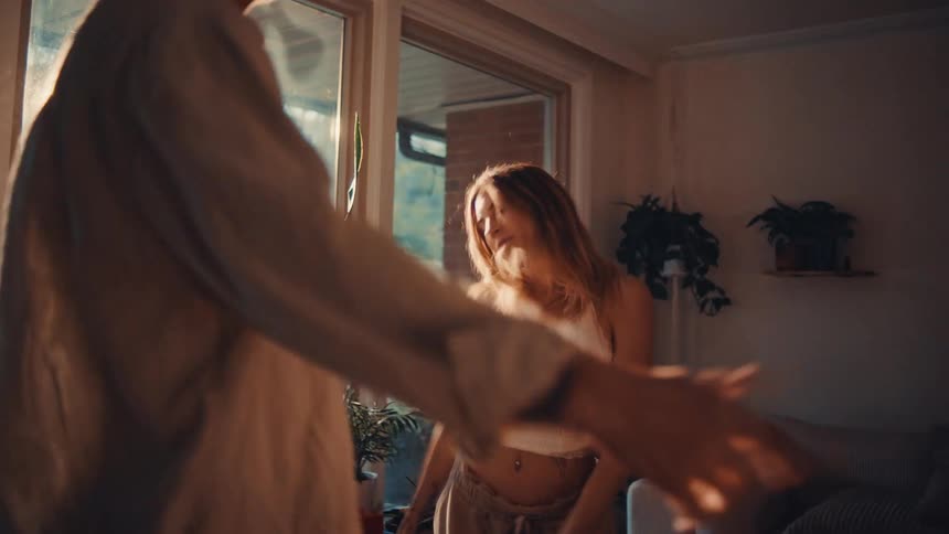 Woman dancing with arms raised beside a man in a sunlit living room