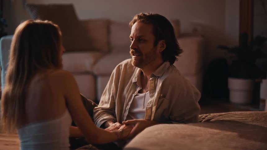 Couple sitting in cozy room sharing affectionate glances and gestures under soft lighting