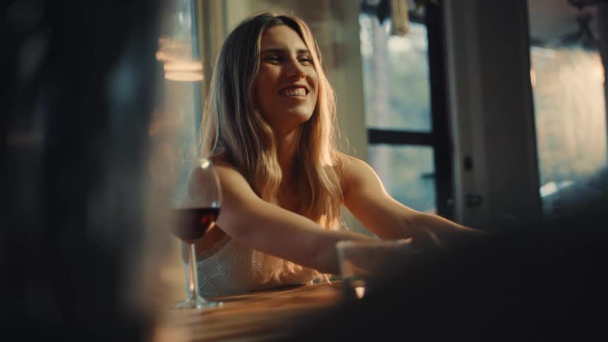 A woman is sitting on a restaurant and talking to someone there is a glass of wine beside her