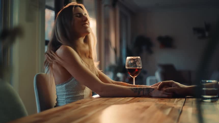 A young woman with long blonde hair sits excitedly in the dining area at midday shaking her body back and forth a glass of red wine is on the table and her hands rest on the table as someone holds them