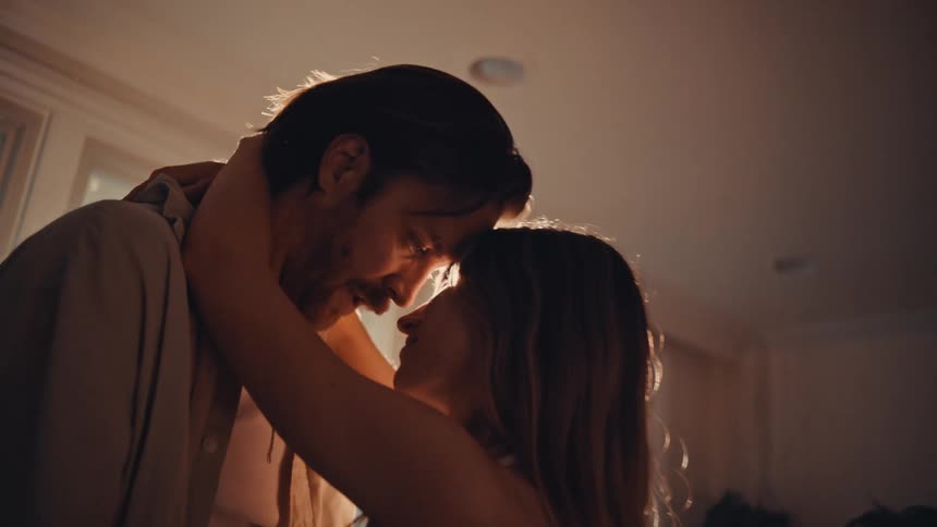 A man and a woman are seen in a closeup embrace their faces nearly touching in a warmly lit intimate settingthe man with a beard and dark hair wears an unbuttoned shirt and the woman with long hair has her arms wrapped around his neck as they