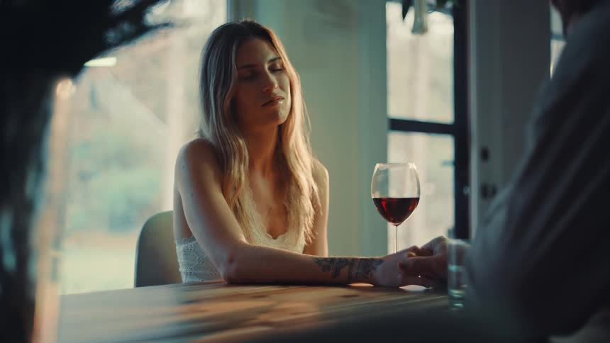 A young woman with blonde hair sits in the dining area at midday looking sad with a glass of red wine in front of her as she holds her spouses hands on the table
