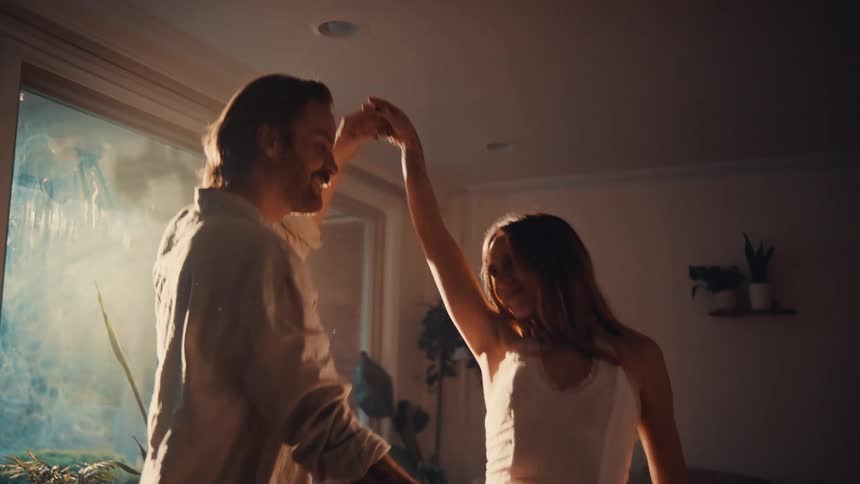 A young couple dance and enjoy themselves in their living room as sunlight streams through the glass window the woman has long blonde hair