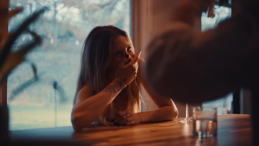 A young woman sits at the dining table at midday with a glass of wine in front of her laughing at the jokes told by the man sitting across from her