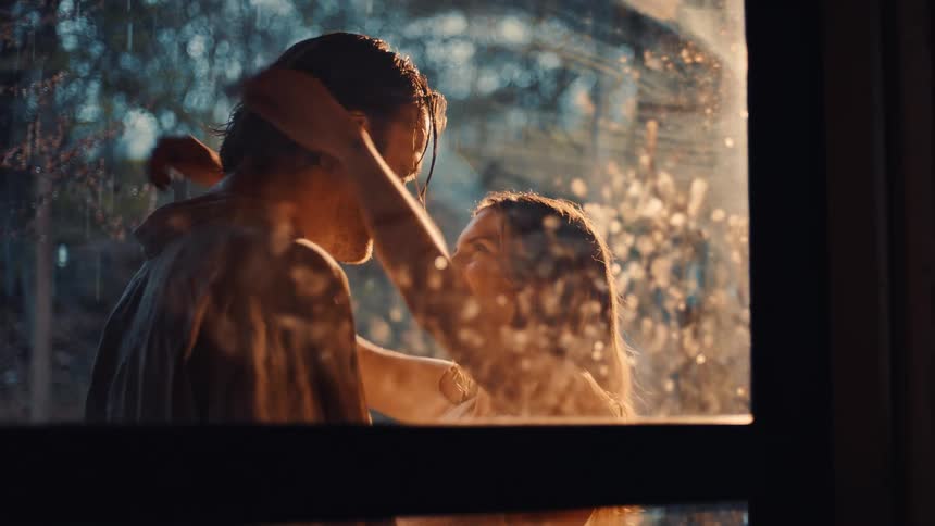 The first clip shows a woman seated at a wooden table with a glass of red wine holding a mans hand and having an intimate conversation in warm dramatic lightingthe second clip shows the same woman and a man embracing tenderly outside a window with