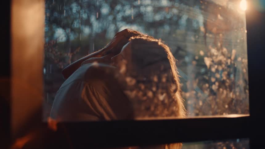 A couple is seen through a rainstreaked window embracing in what appears to be a garden or outdoors setting as rain falls around themthe man and woman pull back slightly to look into each others eyes illuminated by a warm lateday light that catch