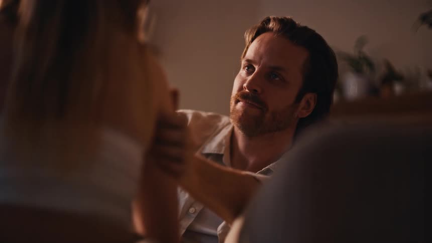 A young couple indoors at midday with the man lying on a couch staring at the young woman with long blonde hair as she rubs his arm on her shoulder