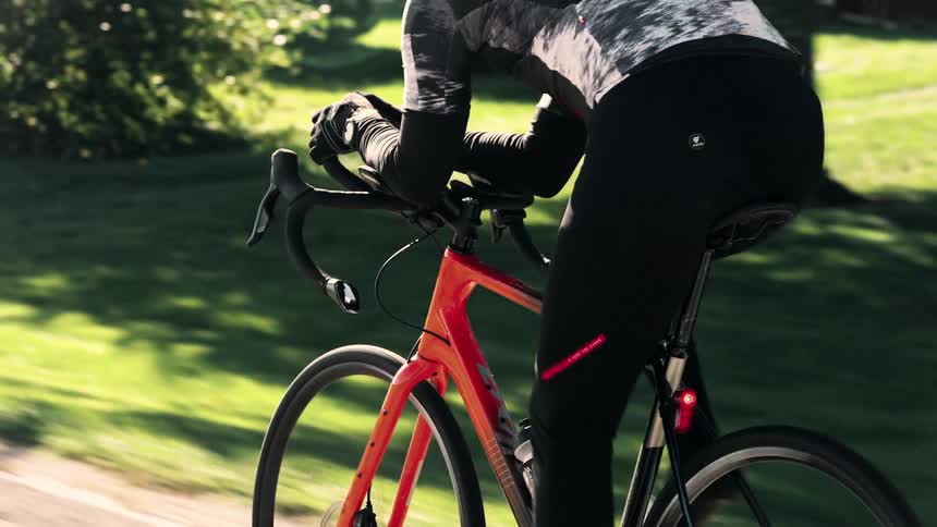 A person cycles on the side of a road lined with trees and grass on a sunny day
