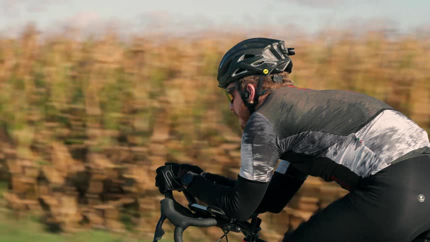 A cyclist leans forward in determination pedaling at speed along a rural road as a golden field blurs past