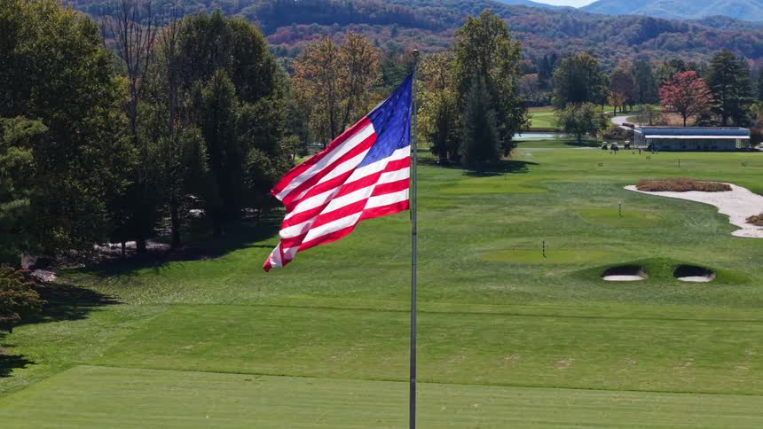 An aerial camera orbits a vibrant american flag waving atop a tall pole revealing a pristine green golf course surrounded by colorful autumn foliage