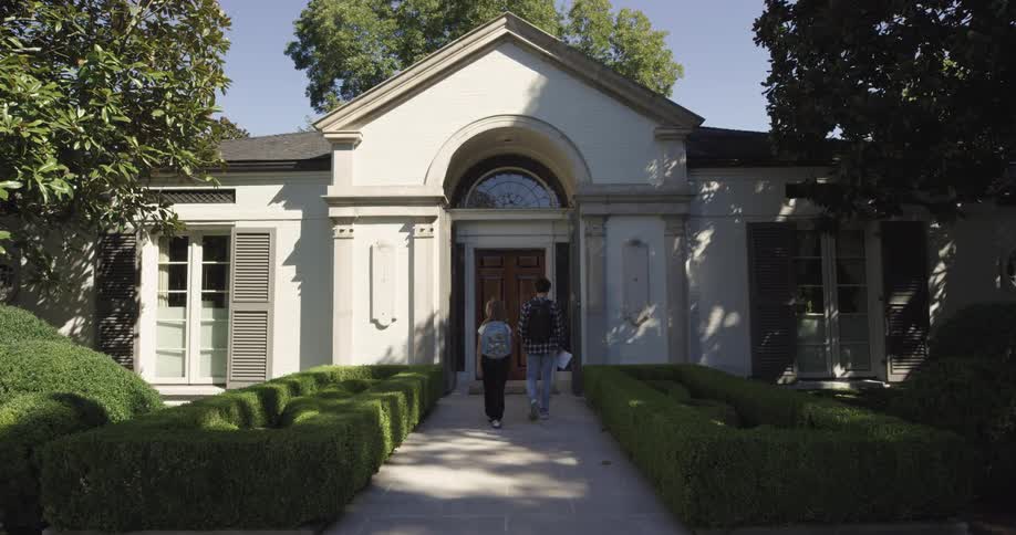 A teenage boy and girl both carrying backpacks walk toward the entrance of a building at midday with a garden on the front lawn