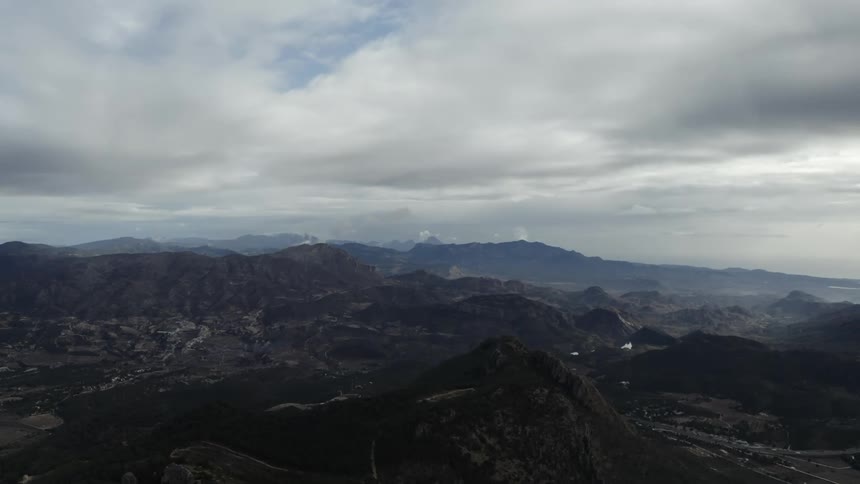A wide aerial view captures a vast dark rugged mountainous landscape stretching to the horizon under a heavily clouded and overcast sky