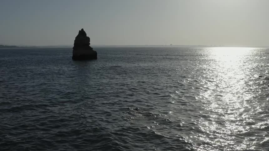 A wide shot frames a tall dark isolated rock pillar rising from the ocean water with intense sunlight glinting across the surface and a hazy sky