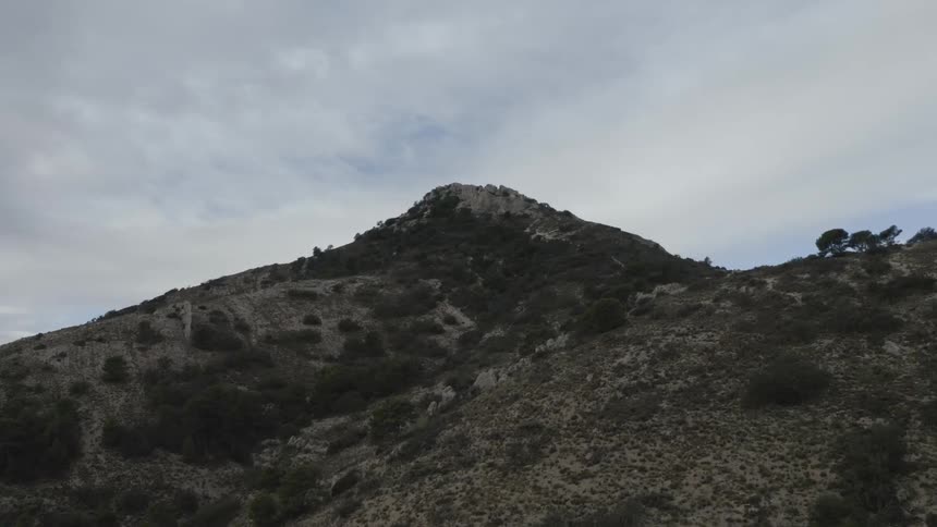 A slow aerial view advances towards a rugged rocky mountain peak rising against a vast overcast sky