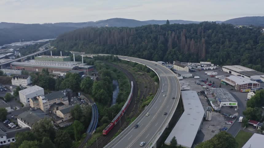 An elevated panning shot moves left to right over a dense valley showing a multilane highway curving over a railway where a red train passes beneath surrounded by industrial buildings and steep forested hills