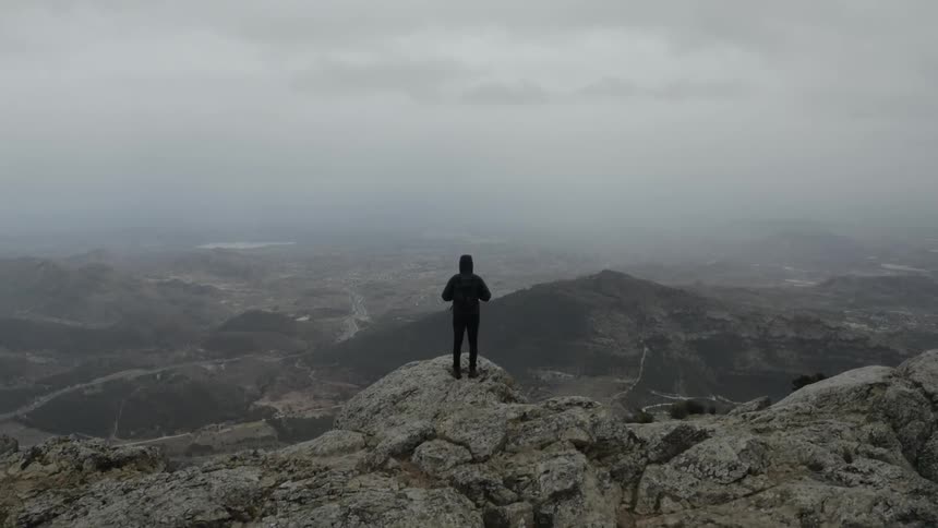 Man stands on top of a rock covered mountain overlooking buildings and roads below