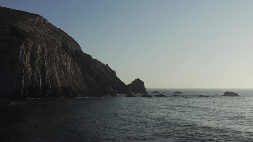 A wide shot frames a rugged towering dark cliff face meeting the ocean water on the left with waves rolling in and splashing against several smaller isolated rock formations under a bright clear blue sky