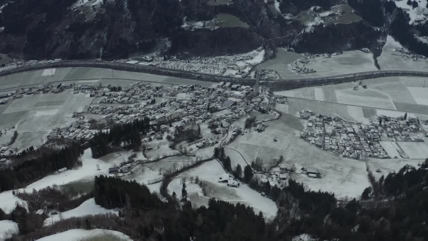 A wide aerial shot shows a dense winter town stretching along a river in a valley surrounded by snow covered fields and dark forested mountains