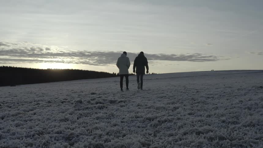 Silhouetted by the low winter sun two men walk together across a wide field blanketed in crisp morning frost