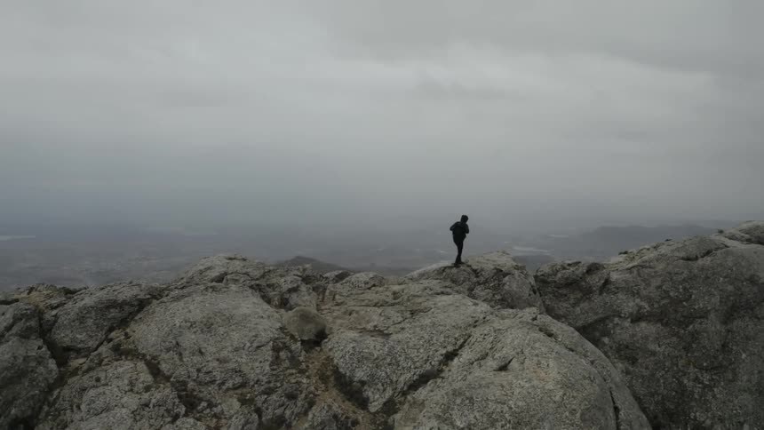 A solitary man wearing a jacket and backpack walks across the jagged rocks of a mountain summit under a heavy overcast sky surveying the expansive hazy landscape below