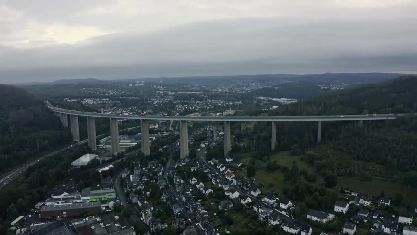 Elevated viaduct spans residential valley supported by concrete pillars amid wooded hills