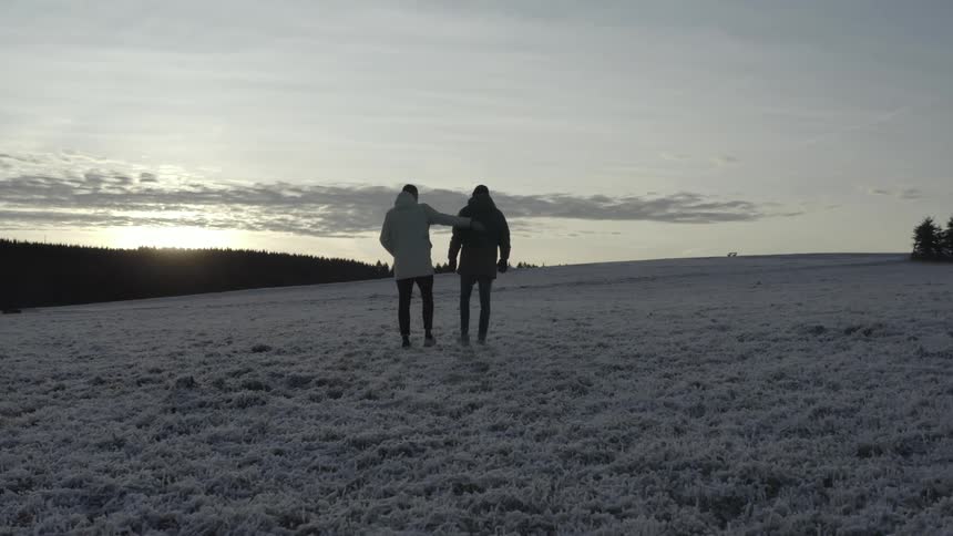 Man touches a friends back as they walk across frozen ground bordering conifer forest while sunlight brightens the horizon