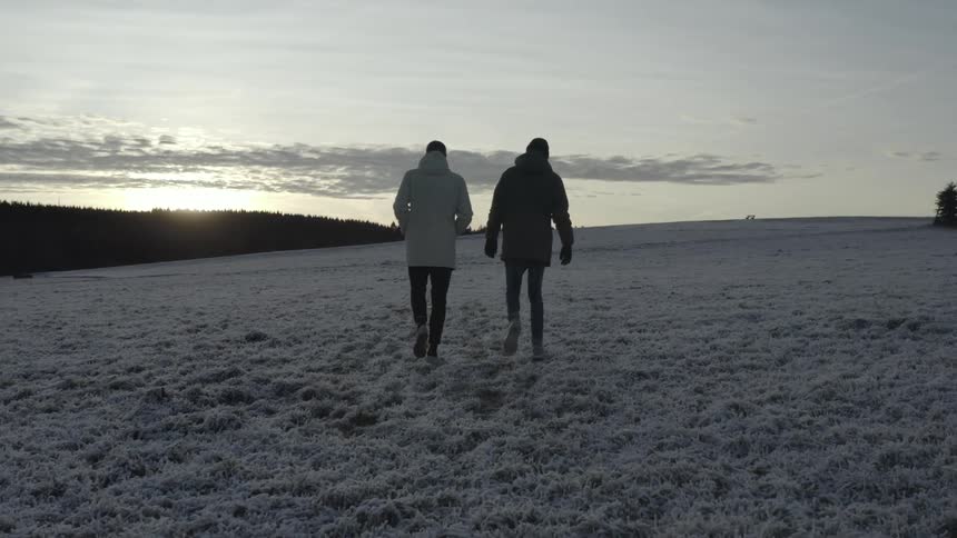 Two young men wearing winter jackets walk in an open area covered in snow at sunset