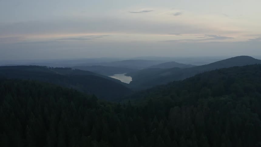 Drifting slowly over a vast dark forest at dusk an aerial view reveals hazy mountain ranges and a glimmering lake in the distance