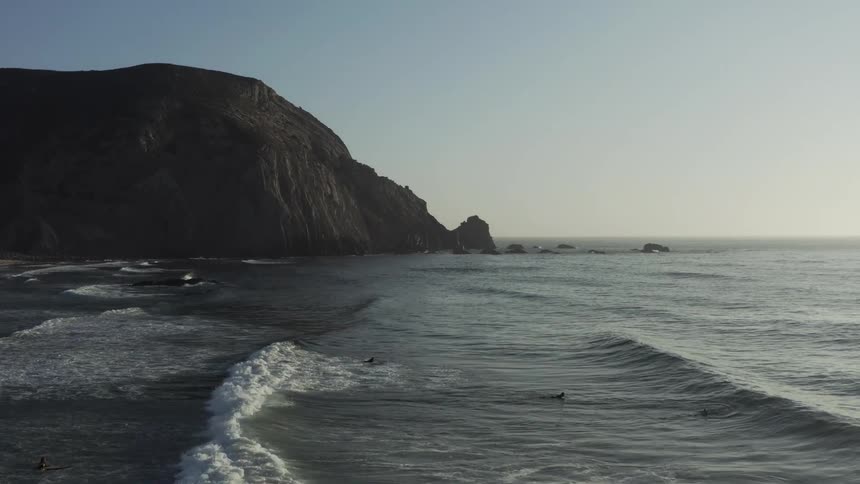 An aerial view drifts over surfers waiting in the calm ocean as gentle waves roll toward a silhouetted cliffside under a pale sky