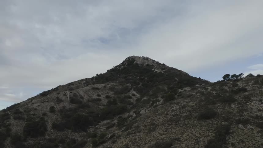 A close view captures a sharp rocky mountain peak with dry scrub covered slopes rising steeply against a cloudy gray sky