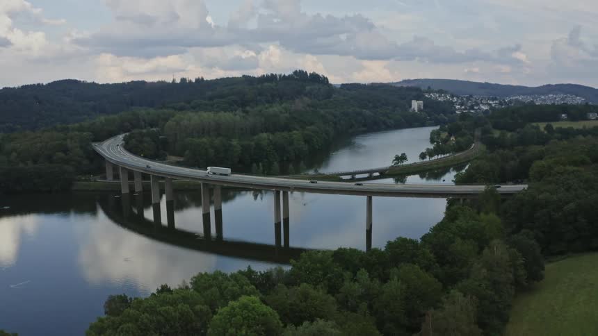 A curved bridge spans a river with a bus and several cars crossing it at midday