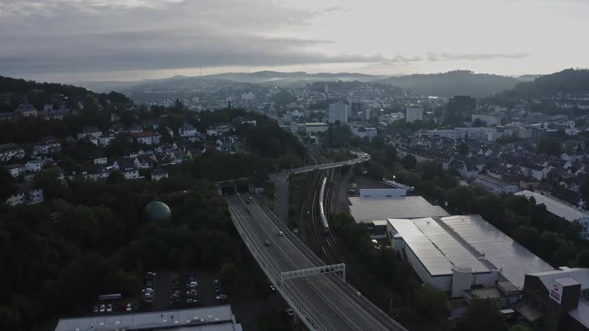 An aerial view captures heavy traffic on a multilevel highway and a train traveling on tracks through a valley city surrounded by forested hills under a cloudy gray sky