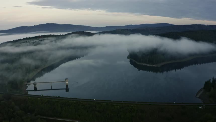 This is an aerial view of a calm reservoir or lake nestled in a heavily forested valley during a misty morning lowhanging fog covers the waters edge and the surrounding dark green wooded hills with a bridge structure visible near the dam