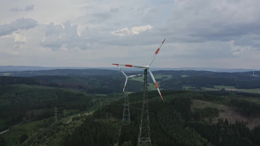 An aerial shot of a hilly forested landscape shows two large wind turbines with white and red blades slowly rotating on tall latticesteel towers as the camera pans slightly over the rolling green hills beneath a thick gray cloudy sky